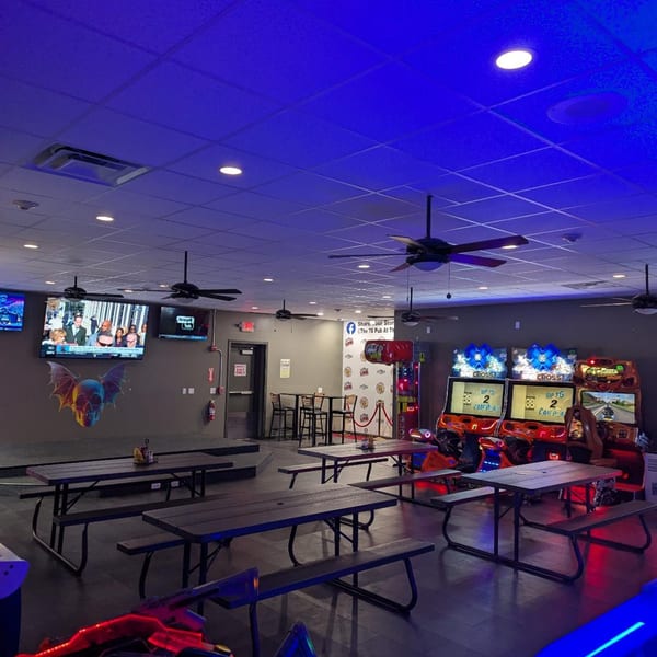 The 78 Pub's interior with picnic tables, arcade games, TVs, and a skull mural under blue lighting.