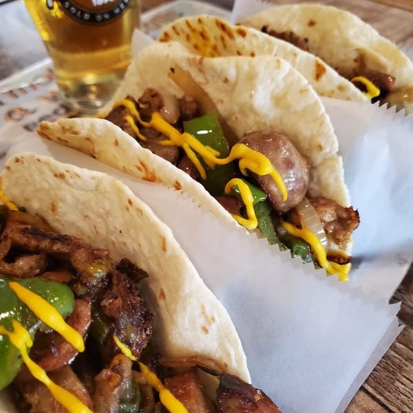 Three Philly cheesesteak tacos with mustard drizzle and a beer in the background.